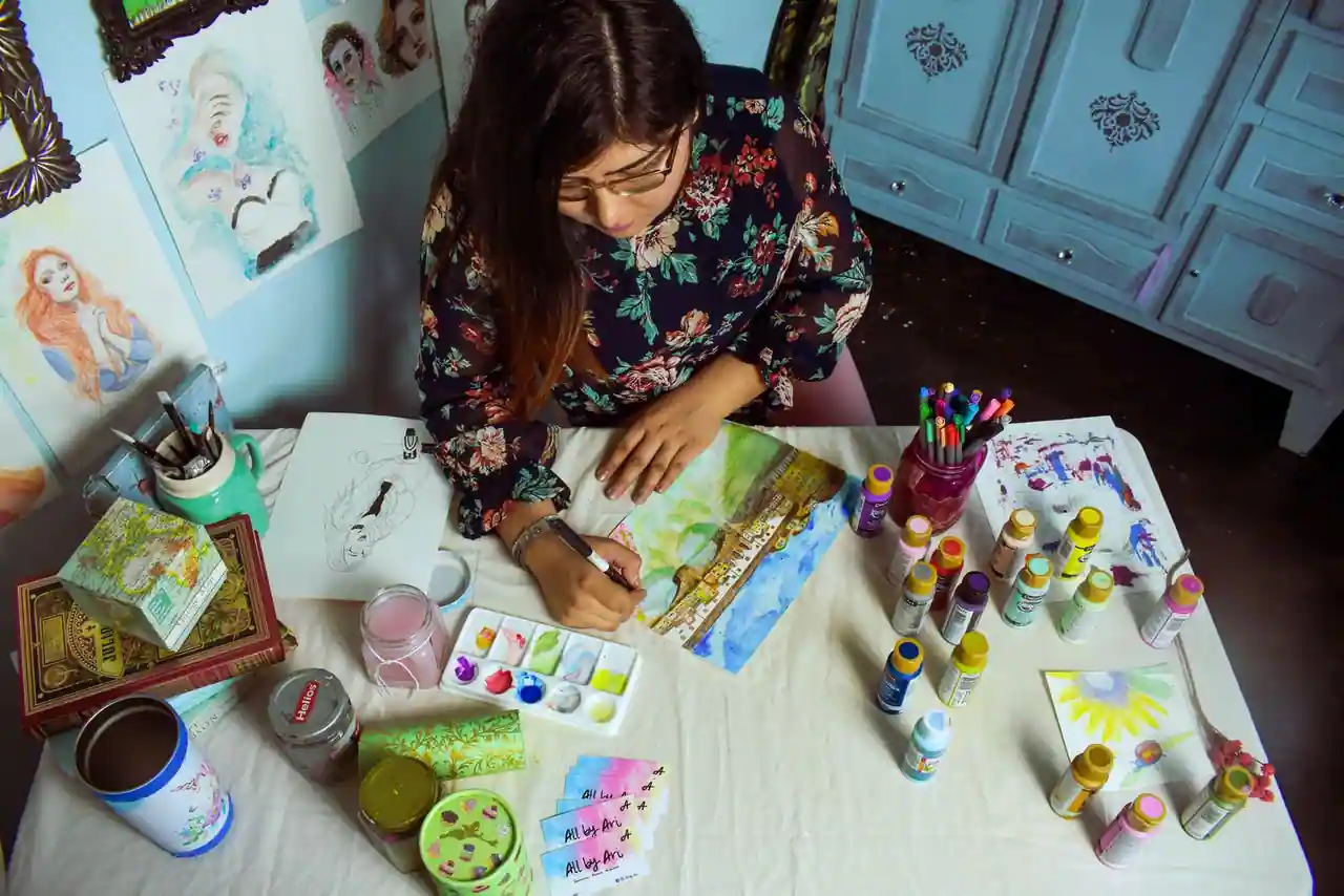 Femme peignant à l'aquarelle à une table couverte de tubes de peinture et de pinceaux, lors d'un loisir créatif.