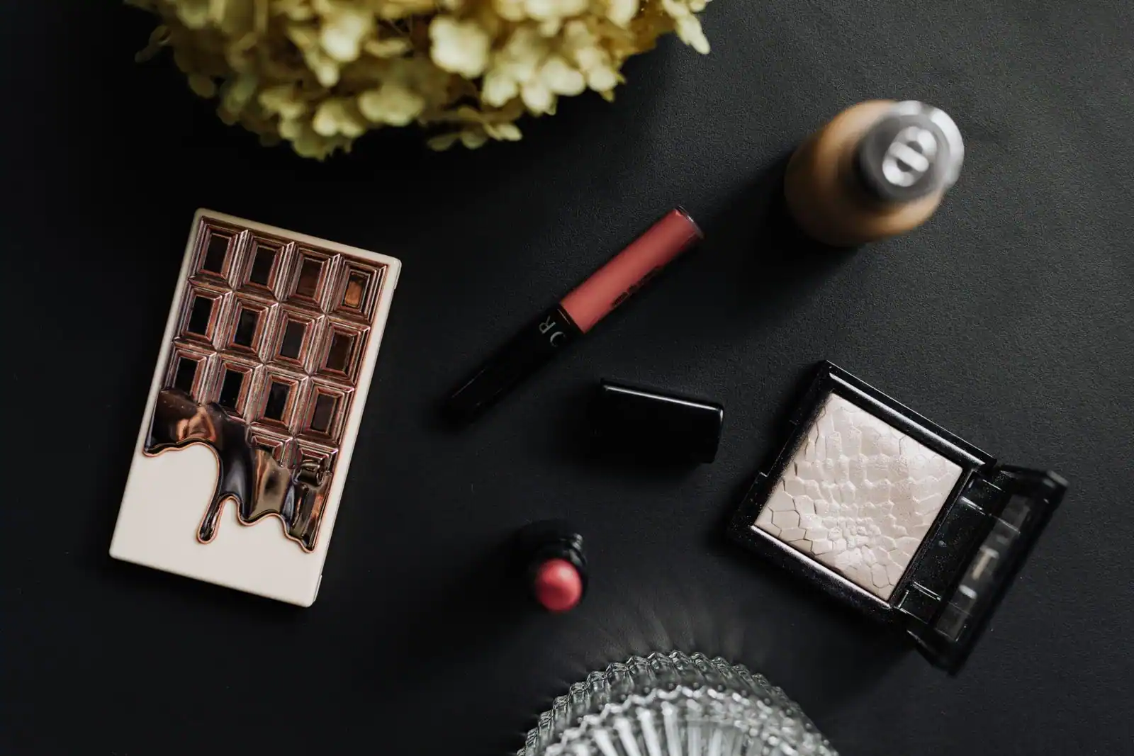 Produits de maquillage — gloss, rouge à lèvres, poudre et palette de fards — disposés sur une surface sombre.