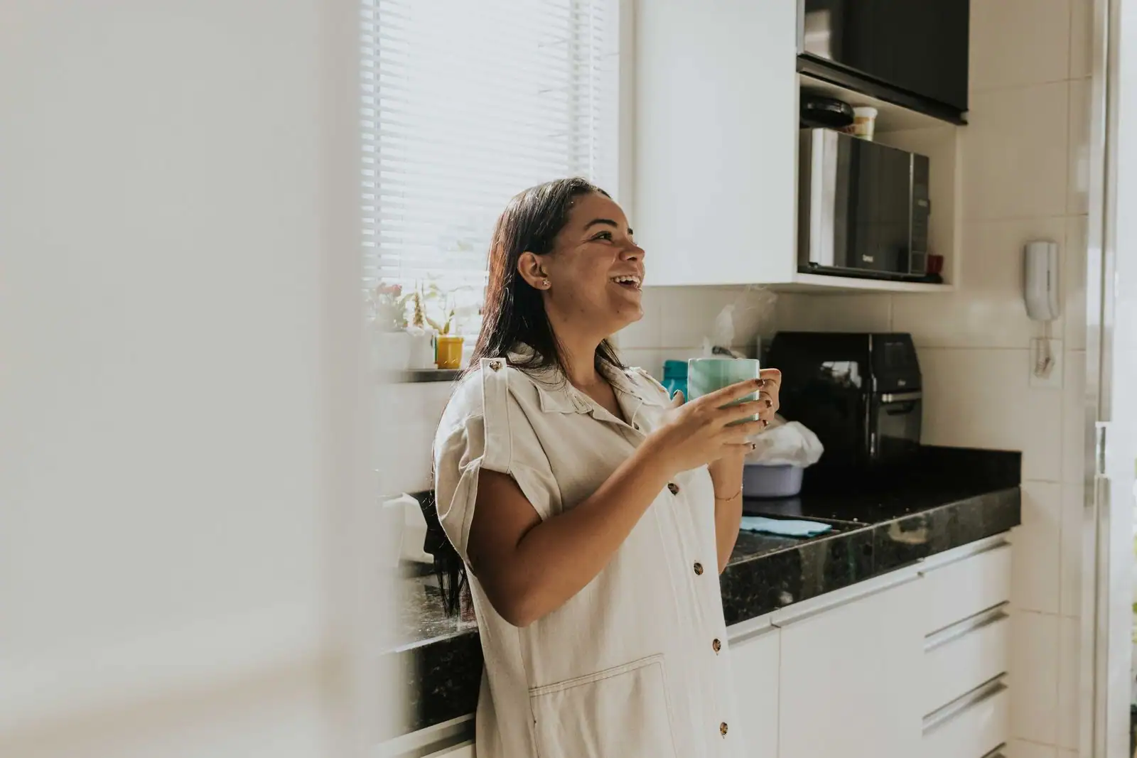 Une femme détendue savoure son café du matin, debout dans une cuisine baignée de lumière.