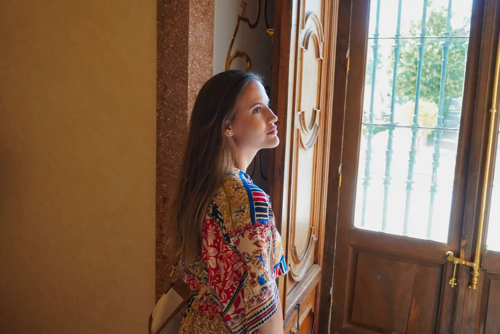 Femme de profil portant une robe à imprimé floral coloré, près d'une porte en bois ensoleillée.