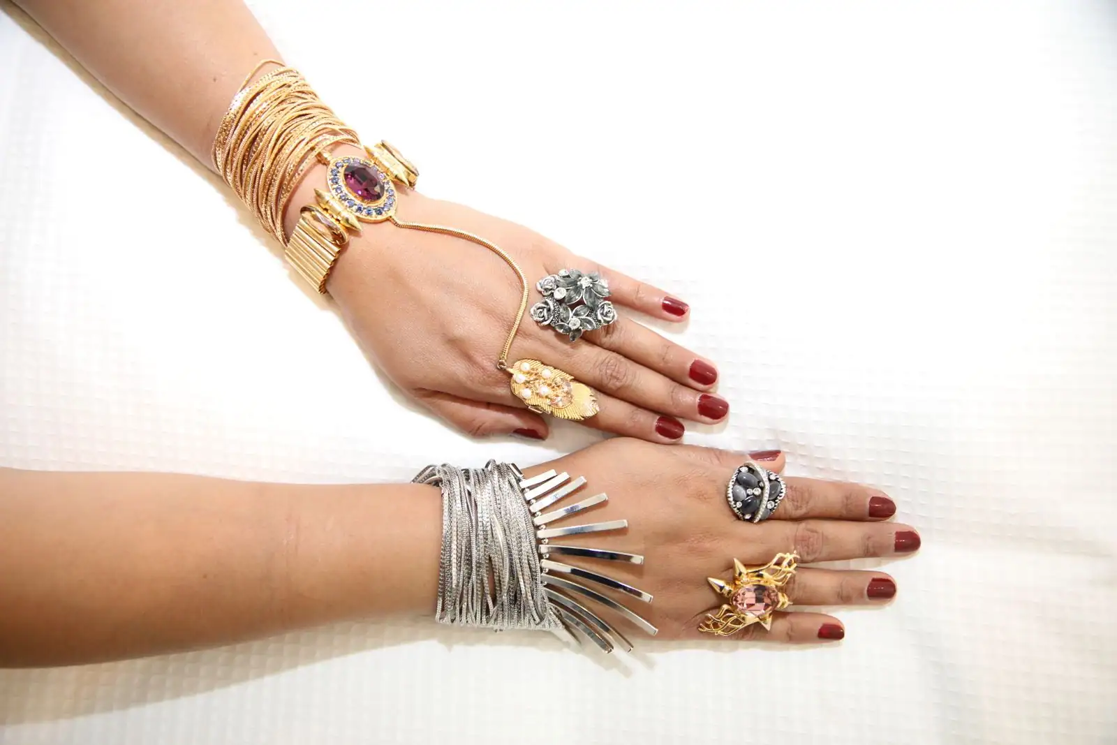 Deux mains portant de nombreux bijoux fantaisie, bracelets dorés et argentés et bagues à pierres colorées.