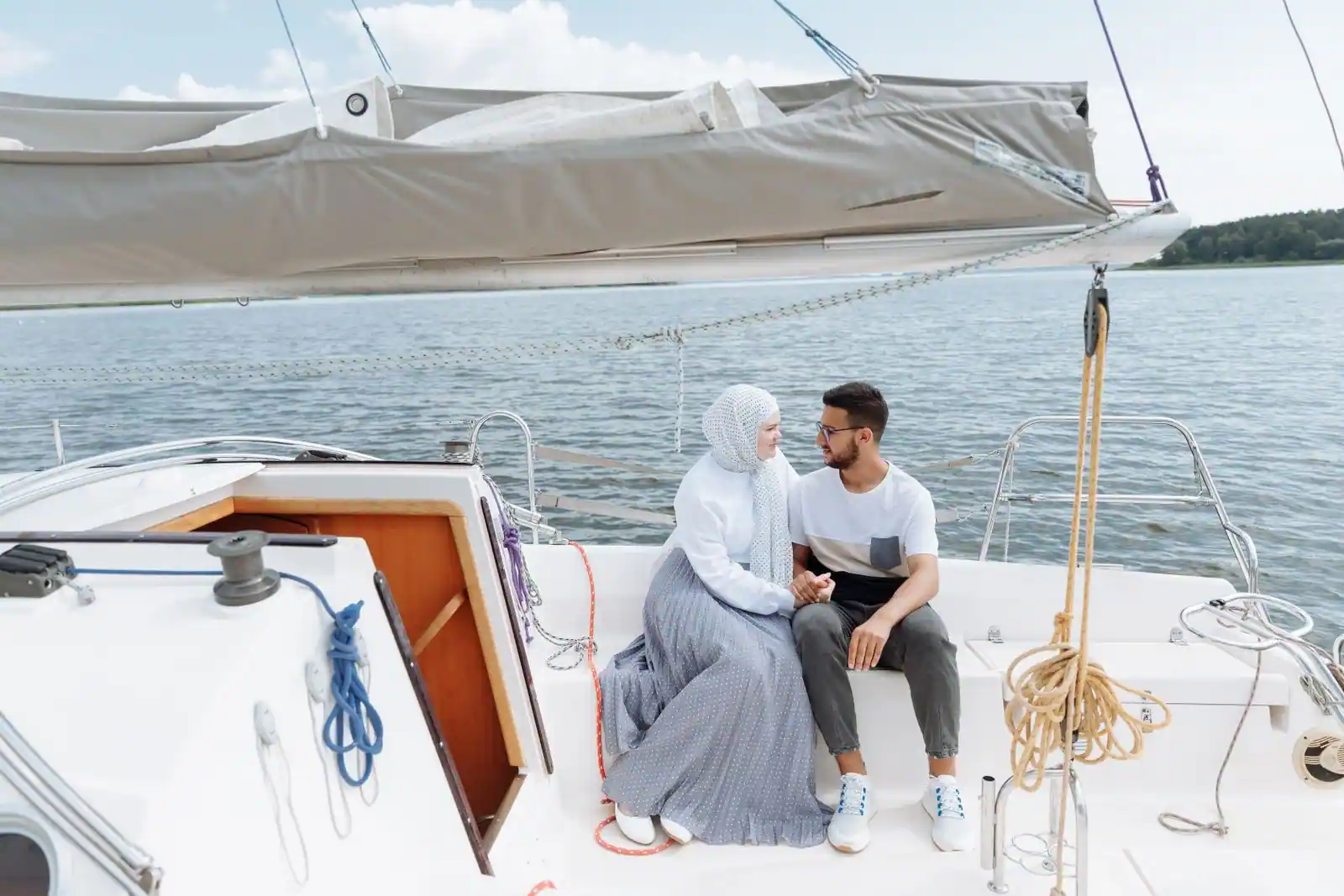 Un couple assis côte à côte sur le pont d'un voilier se regarde tendrement, l'eau calme en arrière-plan.