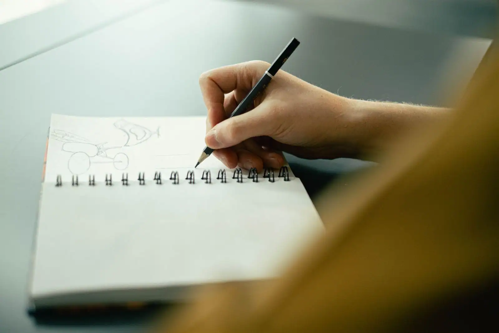 Main esquissant au crayon des croquis simples dans un carnet à spirale posé sur un bureau clair
