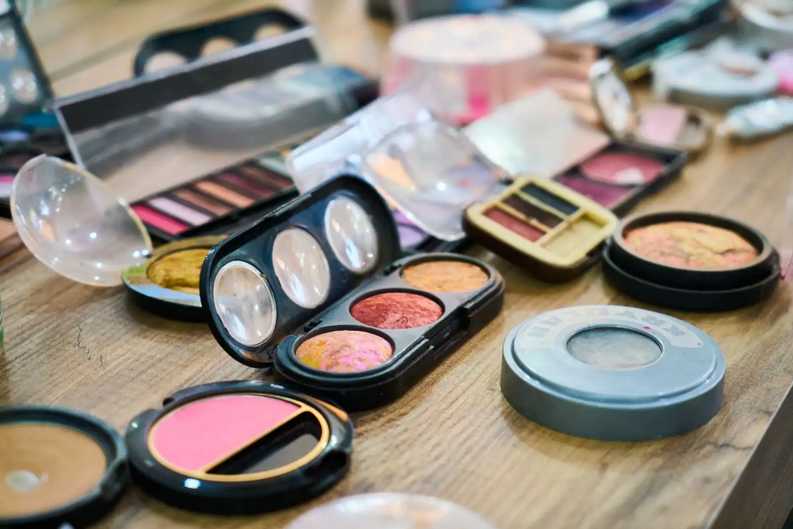 Assortiment de produits de maquillage colorés étalés sur une table en bois : palettes de fards, blushs et poudres.