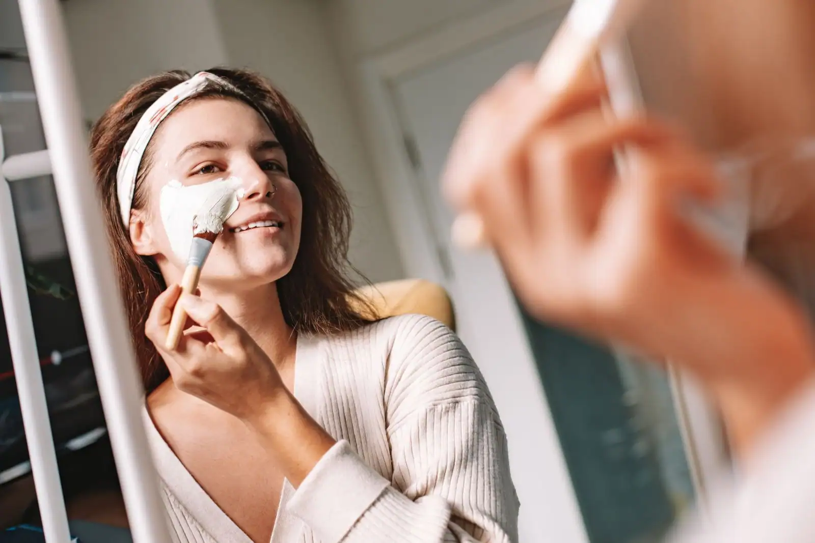 Une femme applique un masque de soin crémeux sur le visage au pinceau, devant son miroir