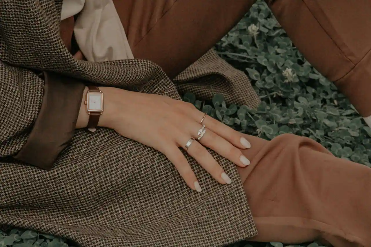 Une montre au boîtier rectangulaire et bracelet en cuir, portée au poignet d'une main aux bagues fines.