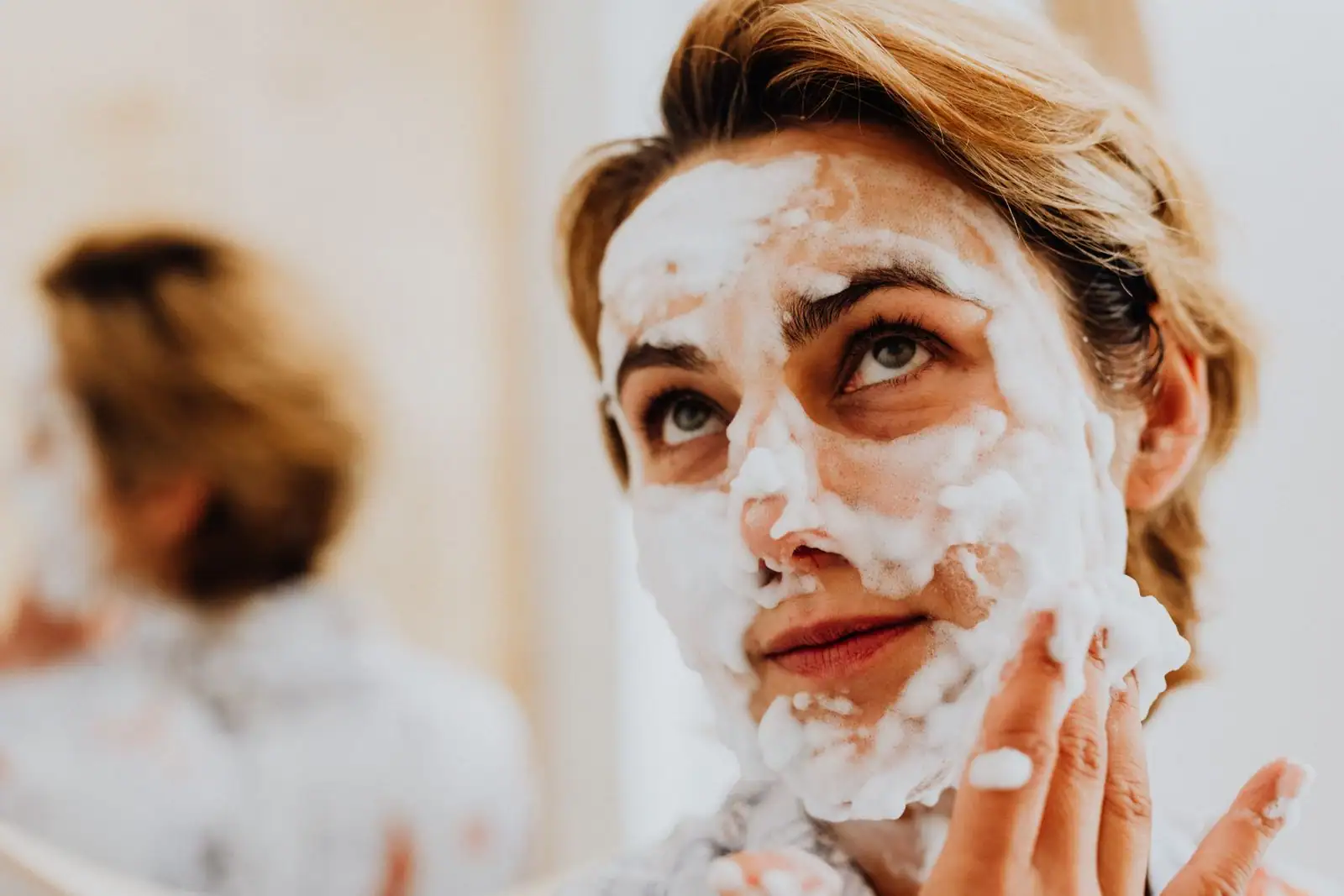 Femme appliquant une mousse nettoyante sur son visage devant un miroir, geste doux du bout des doigts.