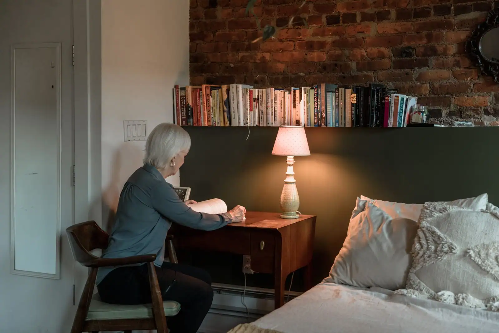 Une femme lit à la lueur d'une lampe dans une chambre chaleureuse en soirée, près d'une étagère de livres.