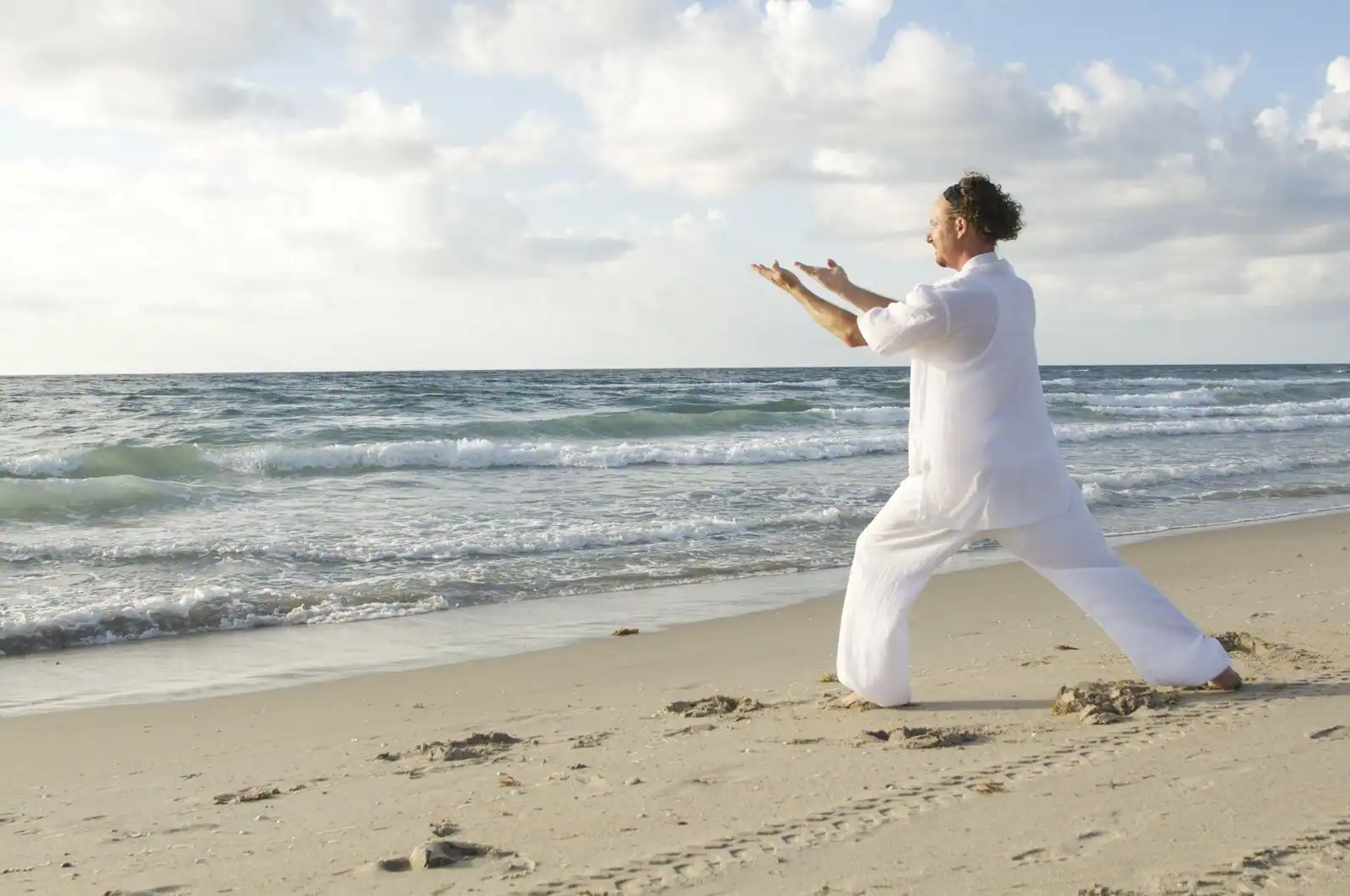 Personne en tenue blanche pratiquant le qi gong au bord de mer, bras tendus dans une lumière douce.