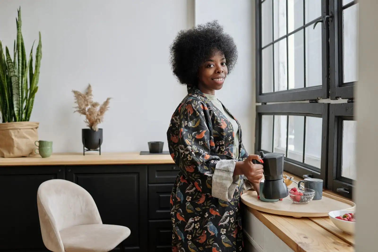 Femme souriante préparant son café du matin près d'une fenêtre lumineuse, ambiance routine matinale apaisée.