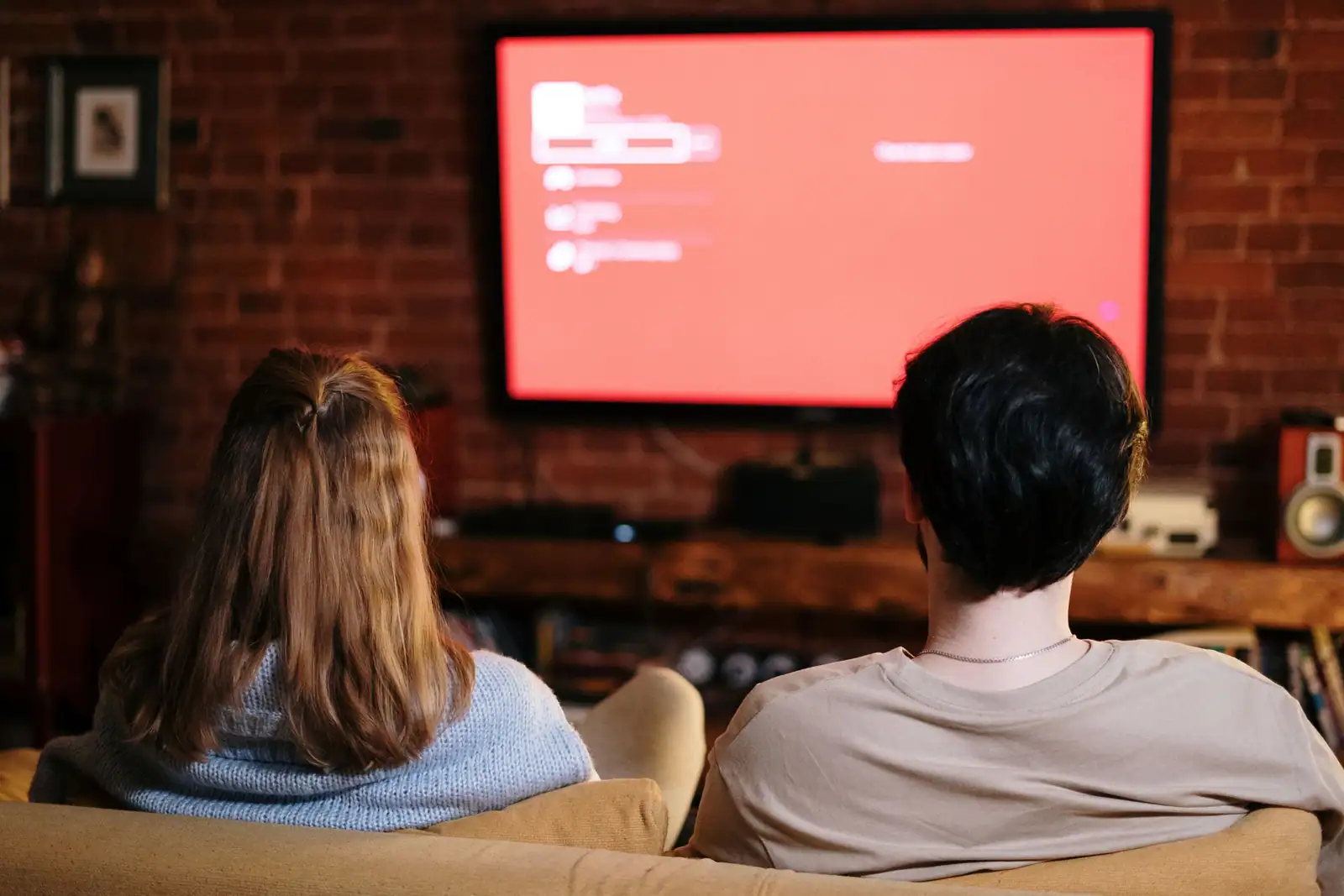 Un couple installé sur un canapé regarde la télévision de dos, dans un salon chaleureux.