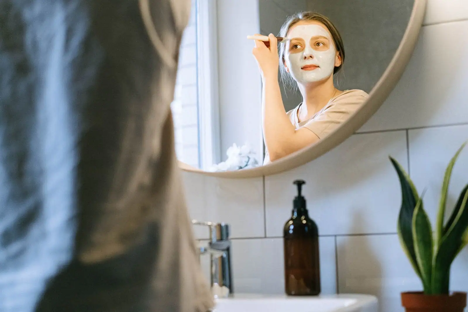 Femme appliquant un masque de soin au pinceau sur son visage, devant un miroir rond de salle de bain