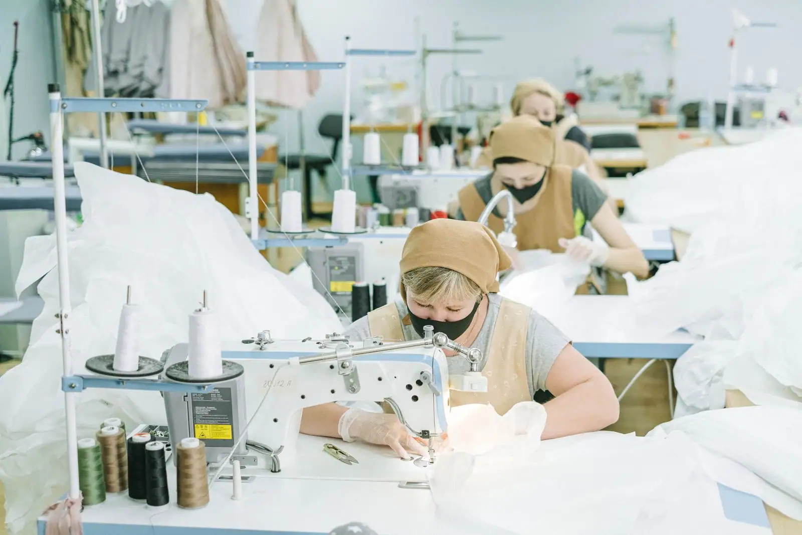 Couturières à leurs machines à coudre industrielles dans un atelier de confection textile, avec bobines de fil et pièces de tissu