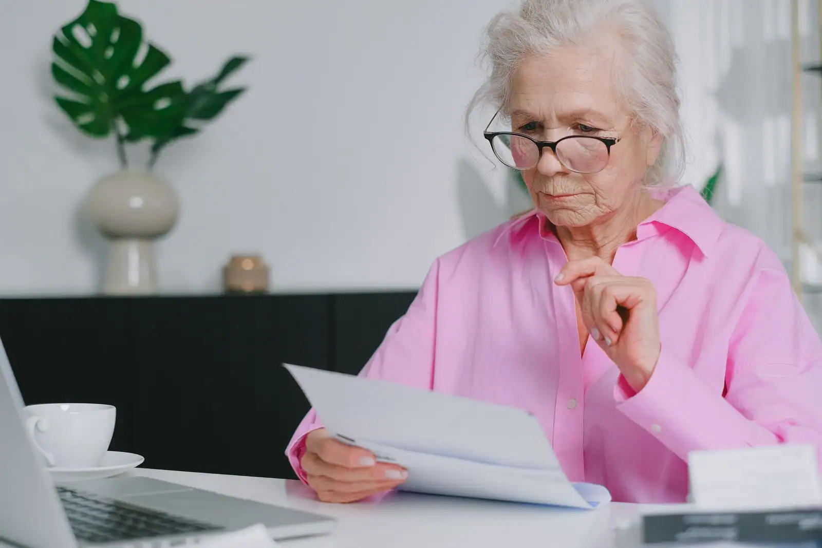 Une femme âgée à lunettes lit attentivement un document administratif à son bureau, chez elle.