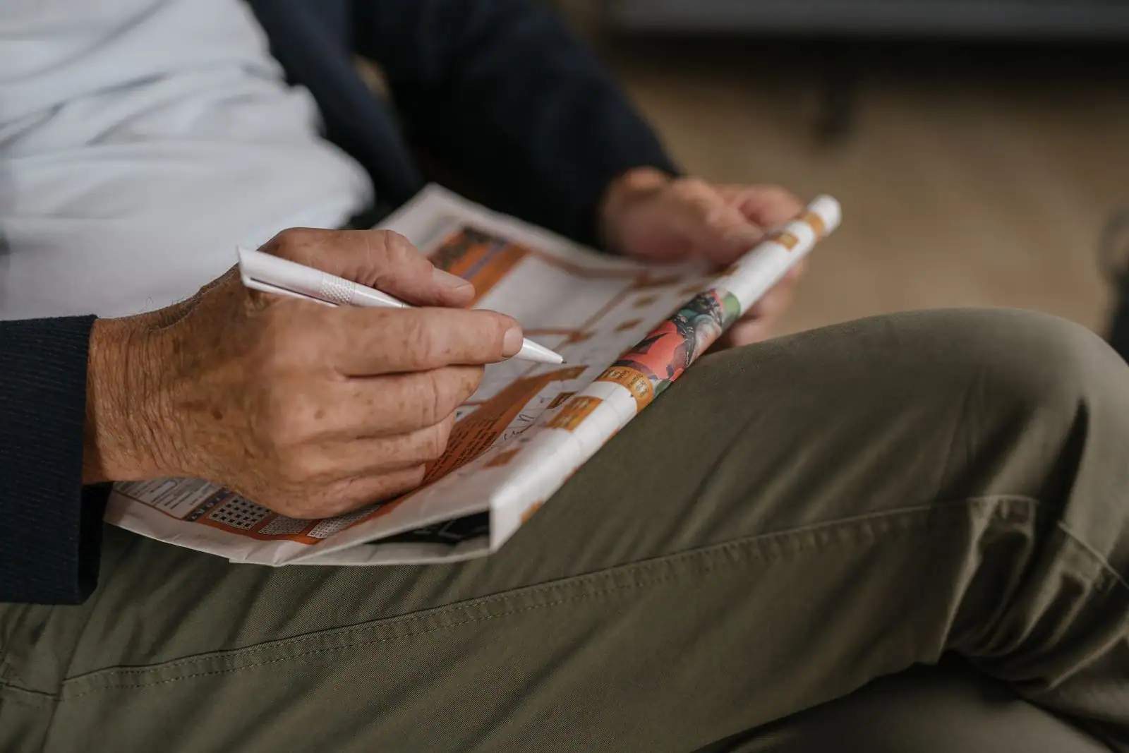 Mains d'une personne remplissant au stylo la grille d'un magazine de jeux plié sur ses genoux.