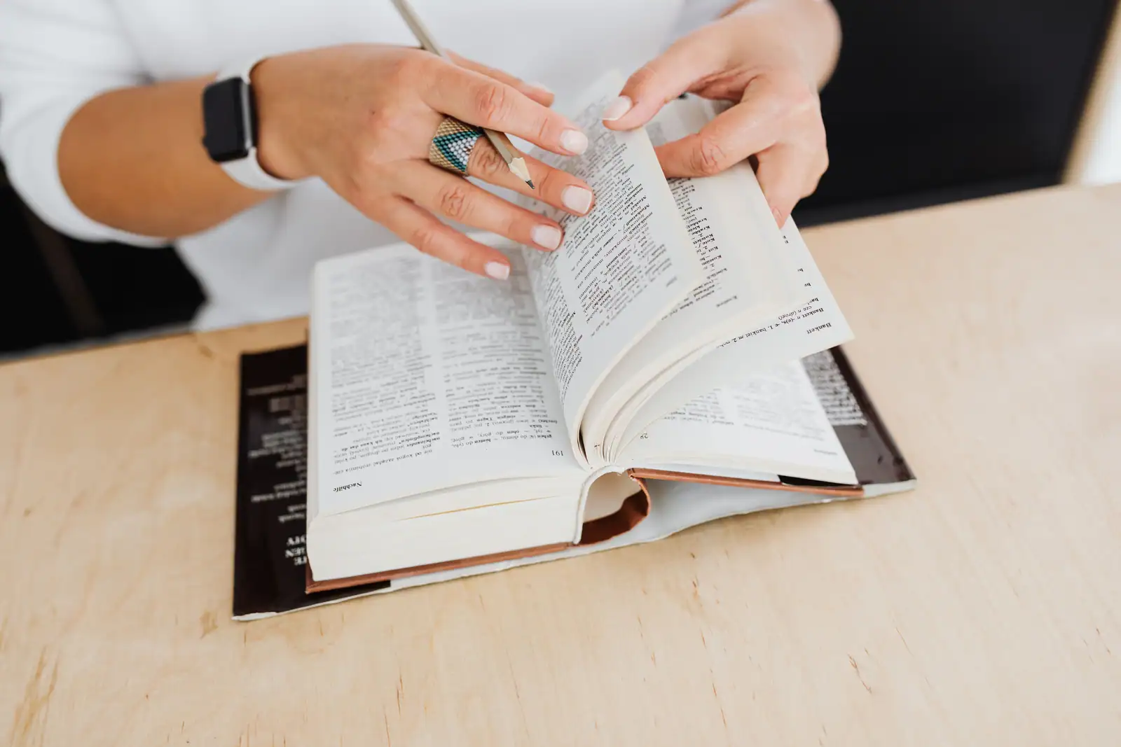 Femme feuilletant un épais dictionnaire relié, un crayon à la main, posé sur un bureau clair