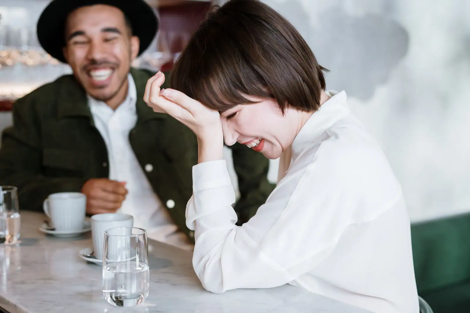 Un homme et une jeune femme éclatent de rire à une table de café, tasses et verres posés devant eux.
