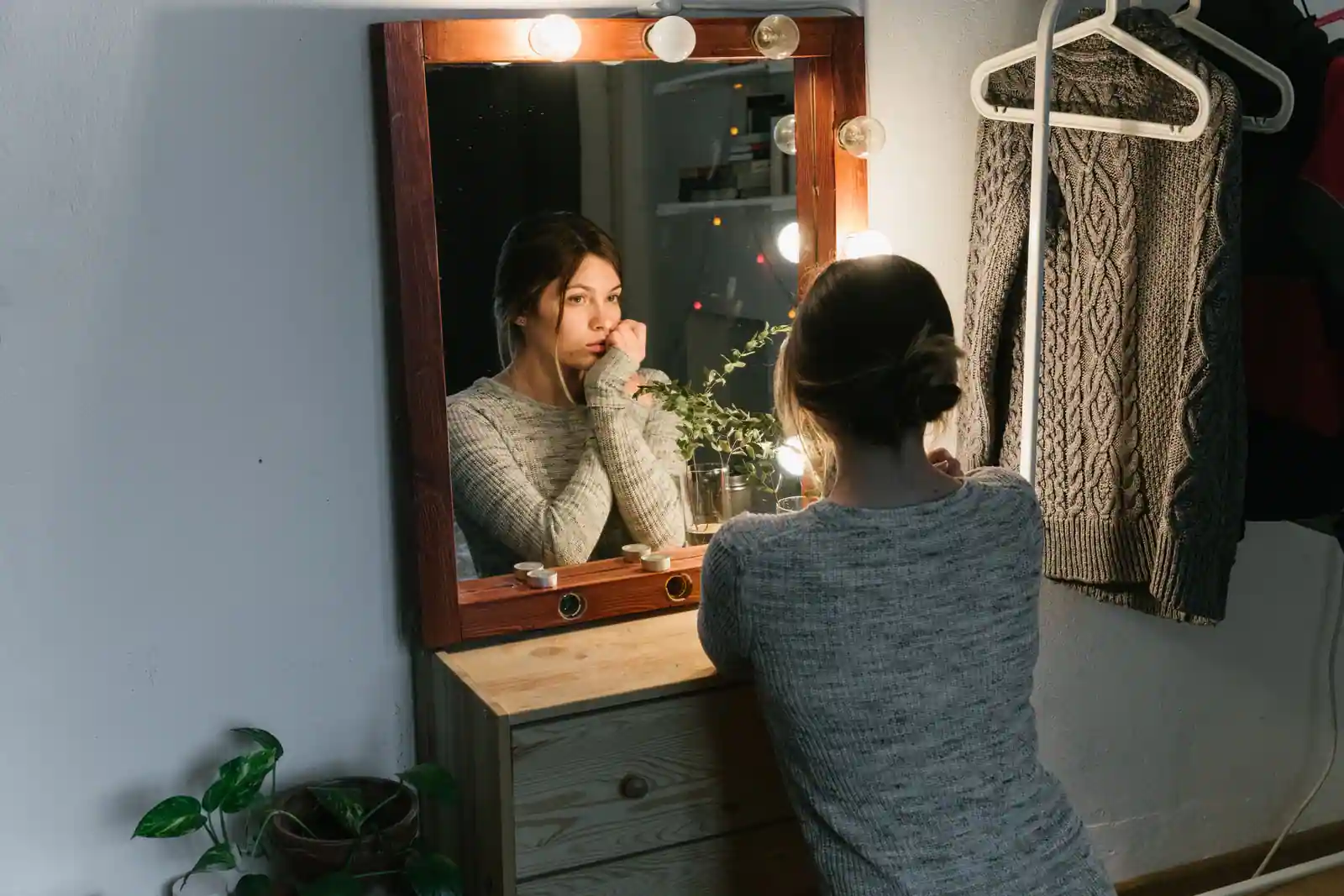 Une femme accoudée à une coiffeuse observe son reflet dans un miroir entouré d'ampoules, l'air pensif