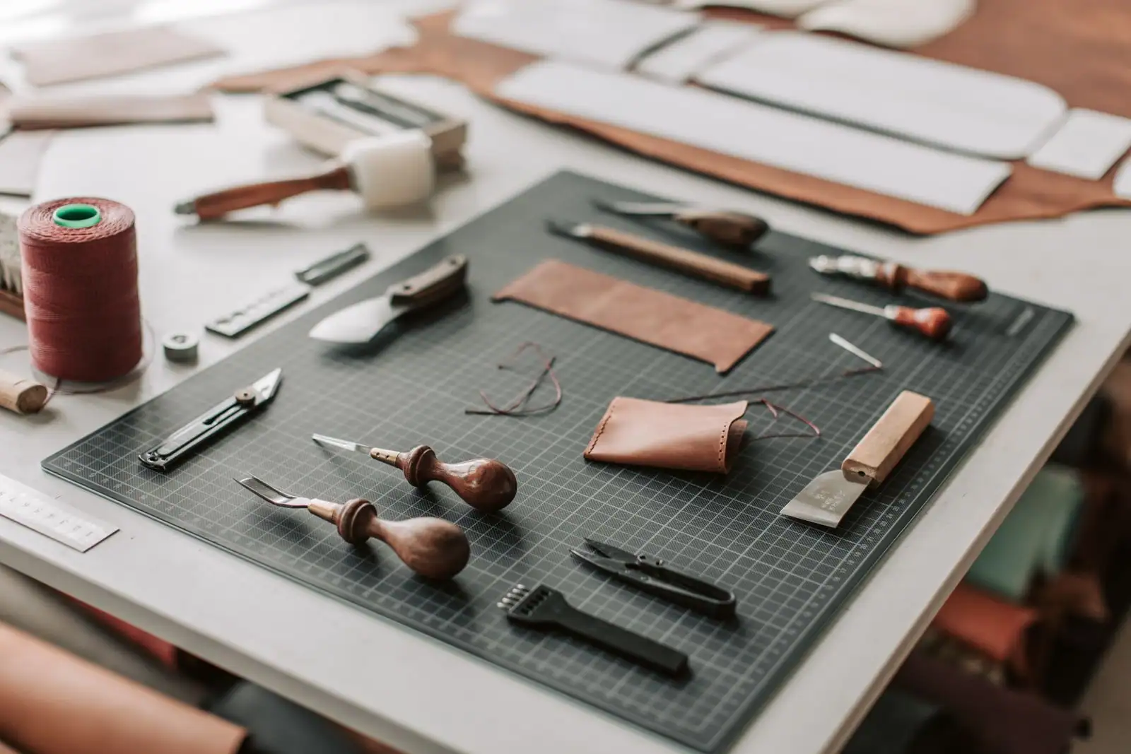 Établi de maroquinerie : outils de coupe, bobine de fil ciré et petit étui en cuir sur un tapis de découpe.