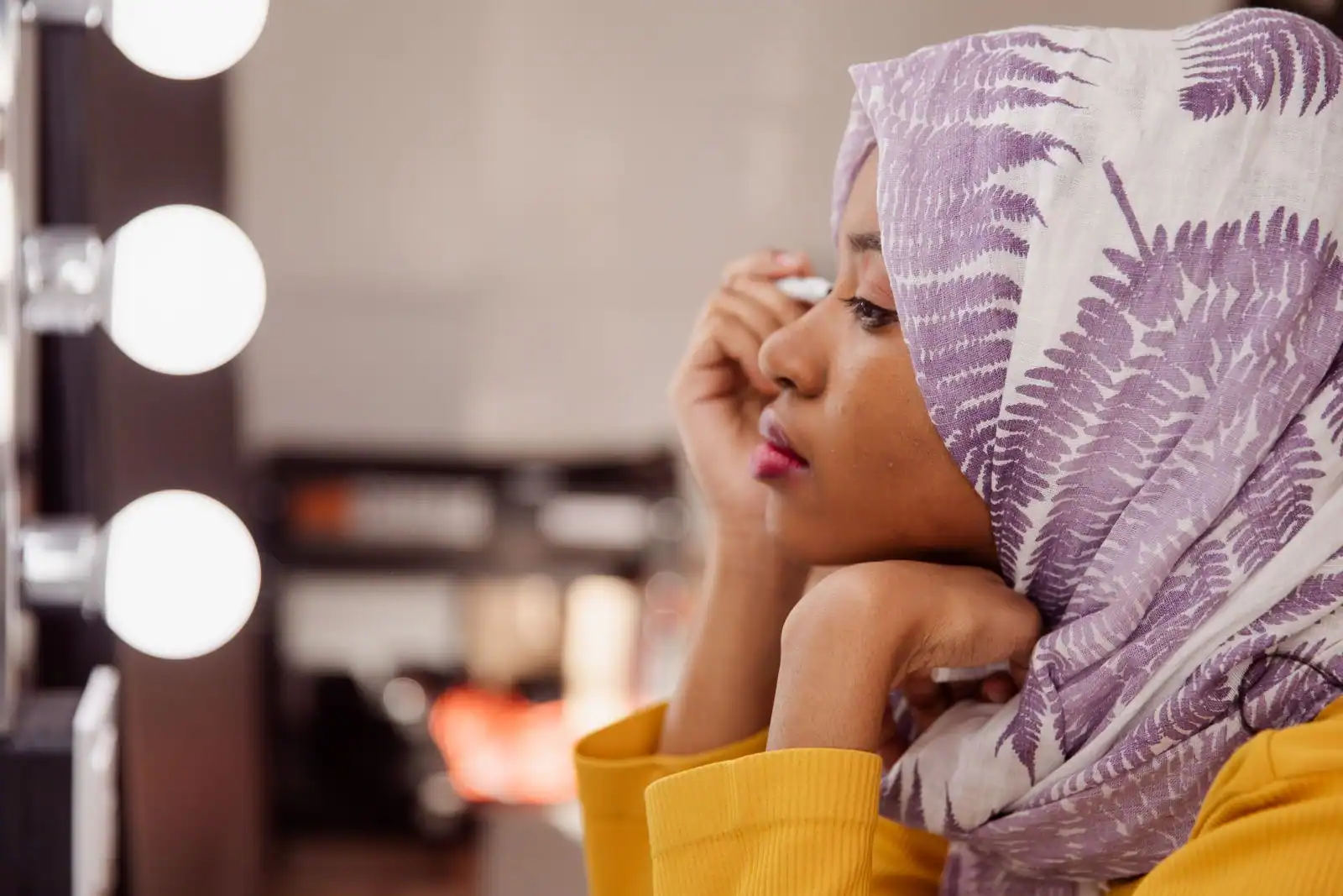Une jeune femme au foulard imprimé applique son maquillage près de l'œil, devant un miroir bordé d'ampoules