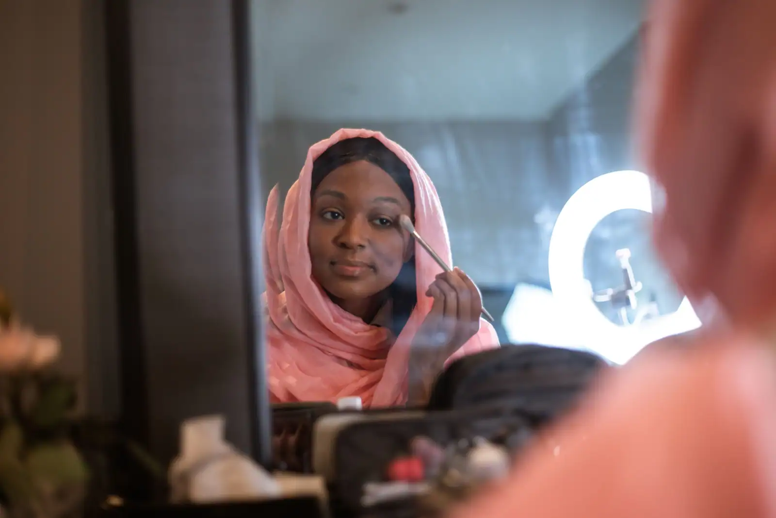 Femme en foulard rose appliquant un fard au pinceau sur la joue, face à un miroir de coiffeuse