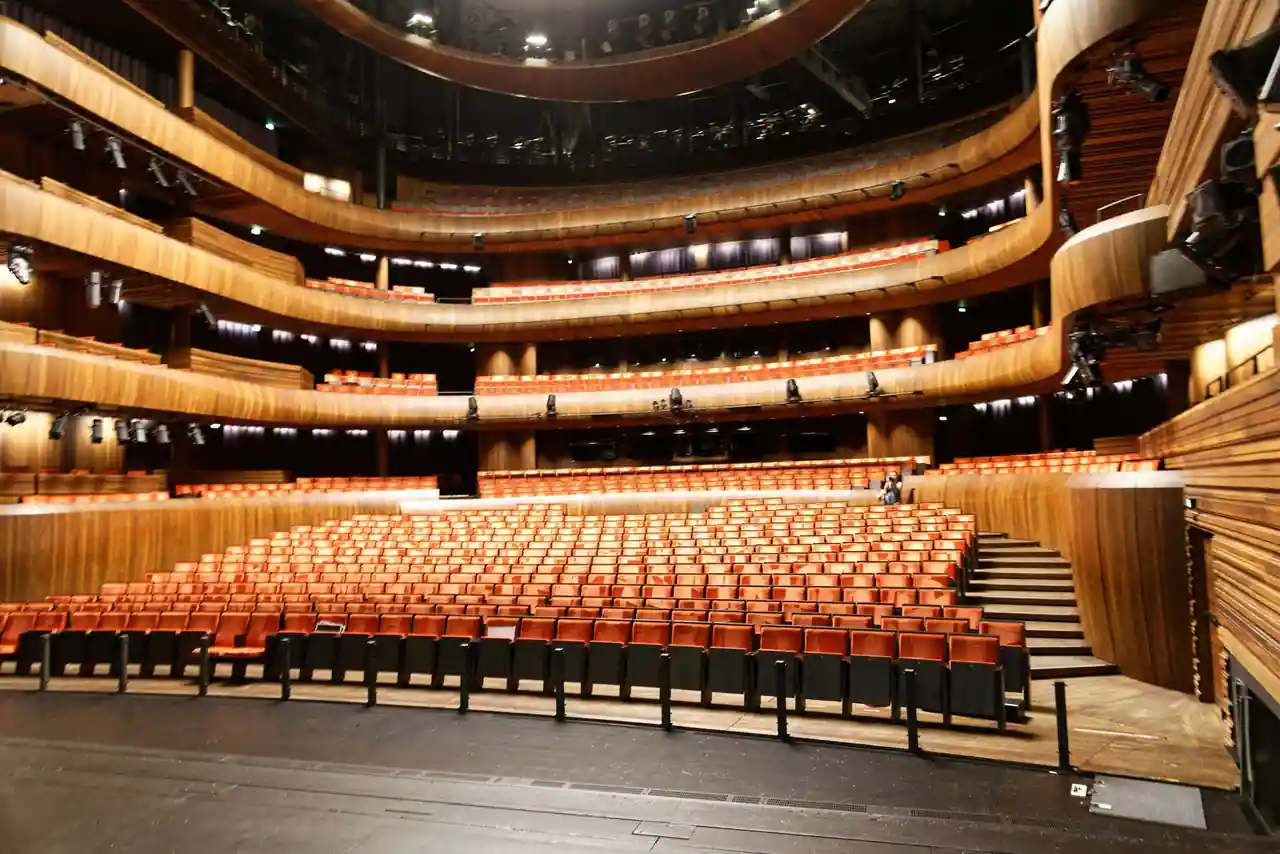 Salle de spectacle vide vue depuis la scène : rangées de fauteuils orangés et balcons en bois