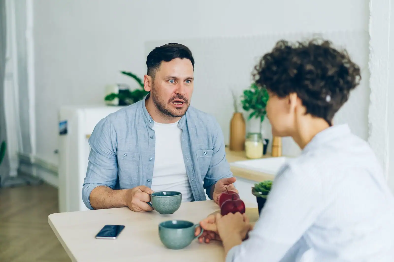Un couple attablé dans une cuisine, tasses de café en main, au milieu d'une conversation tendue.